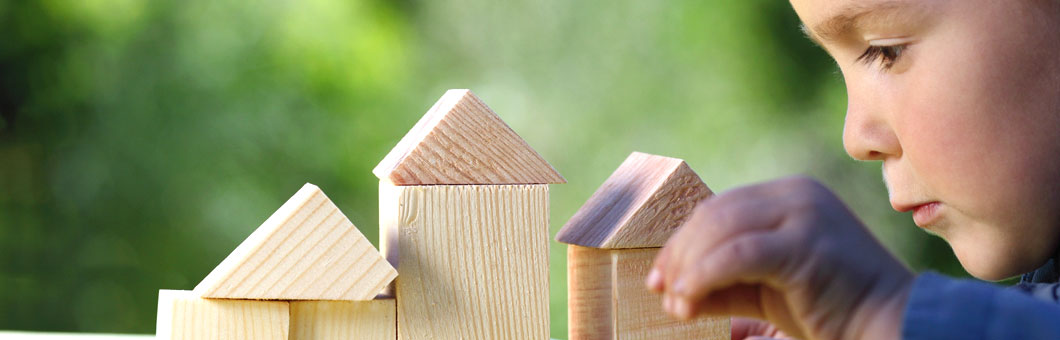 Holzklötze zum Haus zusammengelegt auf einer Tischplatte, rechts im Bild ein Kleinkind, das damit spielt