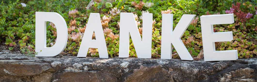 Die Buchstaben D, A, N, K und E stehend auf eine Mauer vor einer Rabatte